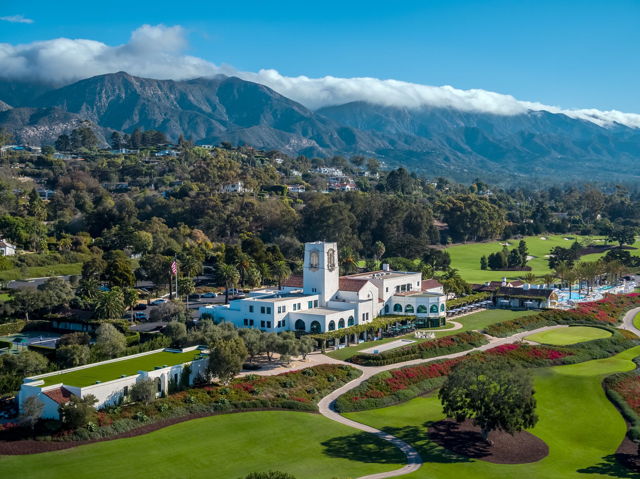 A birds eye view of the Montecito Club, with the large white clubhouse sitting amidst emerald green golf course, trees, and mountains and blue sky in the background.